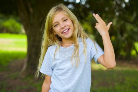 Photo for Caucasian little kid girl wearing white t-shirt standing outdoor in the park foolishness around shoots in temple with fingers makes suicide gesture. Funny model makes finger gun pistol - Royalty Free Image