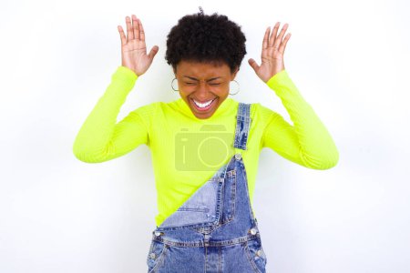 Photo for Young African American woman with short hair wearing denim overall against white wall goes crazy as head goes around feels stressed because of horrible situation - Royalty Free Image