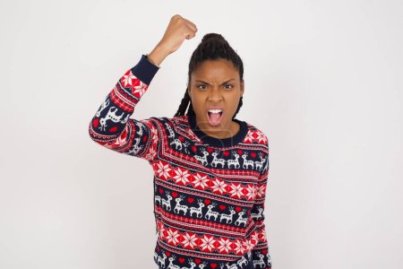 Foto de Mujer afroamericana feroz usando suéter de Navidad contra la pared blanca sosteniendo el puño delante como si estuviera lista para pelear o desafiar, gritando y teniendo expresión agresiva en la cara. - Imagen libre de derechos