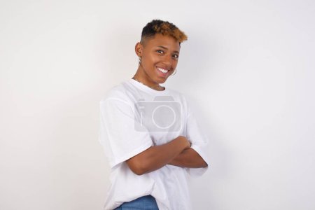 Pretty african-american woman wearing white t-shirt standing against gray wall with arms crossed wearing casual clothes. Looking and smiling at the camera. Confident successful woman.