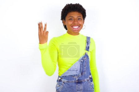 Photo for Young African American woman with short hair wearing denim overall against white wall smiling and looking friendly, showing number three or third with hand forward, counting down - Royalty Free Image