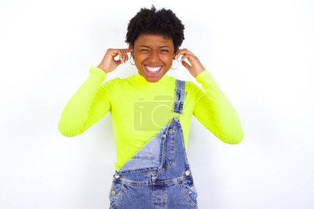 Foto de Feliz joven afroamericana mujer con el pelo corto con denim en general contra la pared blanca ignora la música fuerte y tapones oídos con los dedos pide apagar el sonido - Imagen libre de derechos
