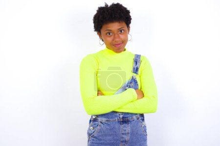 Photo for Waist up shot of  self confident young African American woman with short hair wearing denim overall against white wall has broad smile, crosses arms, happy to meet with colleagues. - Royalty Free Image