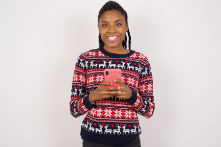 Foto de Sonriente mujer afroamericana usando suéter de Navidad contra la pared blanca amigable y felizmente sosteniendo el teléfono móvil tomando selfie en el espejo. - Imagen libre de derechos