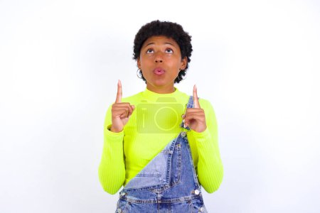 Photo for Young African American woman with short hair wearing denim overall against white wall being amazed and surprised looking and pointing up with fingers showing something strange. - Royalty Free Image