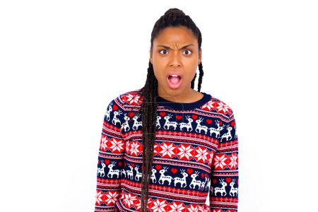 Téléchargez les photos : Expressions faciales expressives. Choqué stupéfait femme afro-américaine portant un pull de Noël contre le mur blanc garde la mâchoire tombée se sent stupéfait de ce qu'il voit de côté. - en image libre de droit