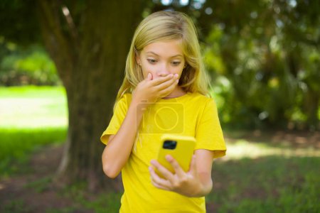 Photo for Caucasian little girl wearing yellow t-shirt standing outdoors being deeply surprised, stares at smartphone display, reads shocking news on website, Omg, its horrible! - Royalty Free Image