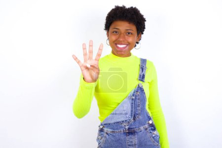 Téléchargez les photos : Jeune afro-américaine aux cheveux courts portant globalement du denim contre un mur blanc montrant et pointant vers le haut avec les doigts numéro quatre tout en souriant confiant et heureux. - en image libre de droit