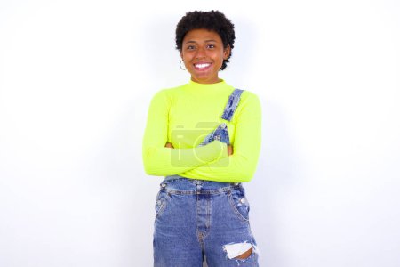 Téléchargez les photos : Jeune afro-américaine aux cheveux courts portant globalement du denim contre un mur blanc heureux souriant et croisé les bras regardant avec confiance la caméra. Personne positive et confiante. - en image libre de droit