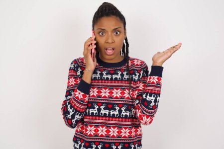 Téléchargez les photos : Femme afro-américaine portant un pull de Noël contre un mur blanc parlant au téléphone stressé avec la main sur le visage, choqué par la honte et le visage surprise, en colère et frustré. Peur et contrariété par erreur. - en image libre de droit