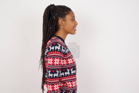 Foto de Perfil de mujer afroamericana sonriente vistiendo suéter navideño contra pared blanca con piel sana, tiene expresión contemplativa, lista para tener paseo al aire libre. - Imagen libre de derechos
