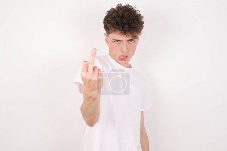 Photo for Young caucasian man with curly hair wearing T-shirt white background shows middle finger bad sign asks not to bother. Provocation and rude attitude. - Royalty Free Image