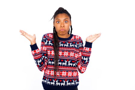 Foto de Mujer afroamericana desconcertada y despistada usando suéter de Navidad contra la pared blanca con los brazos extendidos, encogiéndose de hombros, diciendo: a quién le importa, y qué, no sé. - Imagen libre de derechos