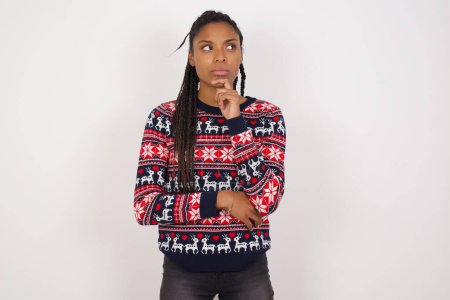 Téléchargez les photos : Prise de vue de contemplative femme afro-américaine réfléchie portant un pull de Noël contre un mur blanc garde la main sous le menton, regarde vers le haut. - en image libre de droit