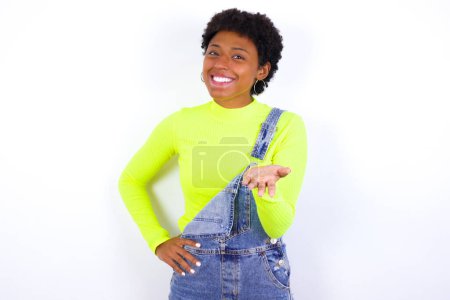 Photo for Young African American woman with short hair wearing denim overall against white smiling friendly offering something with open hand or handshake as greeting and welcoming. Successful business. - Royalty Free Image