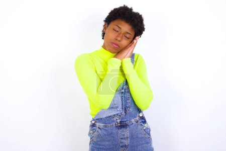 Foto de Relájate y duerme. Mujer afroamericana cansada con el pelo corto usando denim en general contra la pared blanca con los ojos cerrados apoyados en las palmas haciendo el gesto de dormir. - Imagen libre de derechos