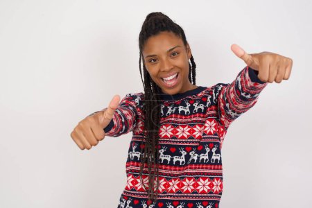 Téléchargez les photos : Femme afro-américaine portant un pull de Noël contre un mur blanc faisant un geste positif avec les pouces levés souriant et heureux pour le succès. En regardant la caméra, geste gagnant. - en image libre de droit