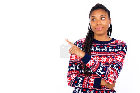 Photo for Portrait of African American woman wearing Christmas sweater against white wall posing on camera with tricky look, presenting product with index finger. Advertisement concept. - Royalty Free Image
