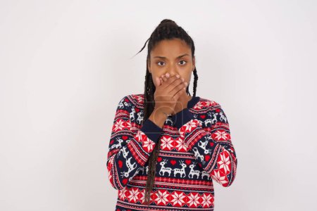 Téléchargez les photos : Femme afro-américaine bouleversée portant un pull de Noël contre un mur blanc couvrant sa bouche avec les deux paumes pour empêcher les cris, après avoir vu ou entendu quelque chose de mauvais. - en image libre de droit