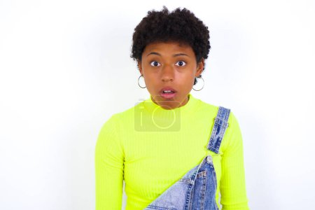 Foto de Joven mujer afroamericana conmocionada con el pelo corto usando denim en general contra las miradas de pared blanca ojos con micrófonos mantiene la boca abierta ha sorprendido a la expresión. Concepto de Omg - Imagen libre de derechos
