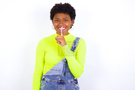 Photo for Smiling African American woman with short hair wearing denim overall against white wall makes shush gesture, holds fore finger over lips hides secret. Be mute, please. - Royalty Free Image