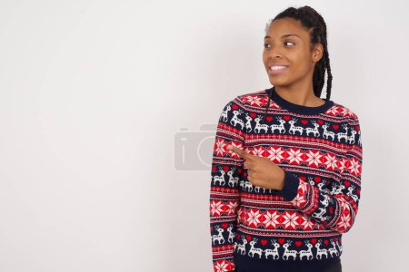 Foto de Feliz alegre sonriente mujer afroamericana usando suéter de Navidad contra la pared blanca mirando y señalando a un lado con la mano. Copiar espacio y concepto de publicidad. - Imagen libre de derechos