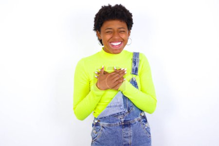 Foto de Mujer afroamericana con el pelo corto con denim en general contra la pared blanca expresa happines, ríe agradablemente, mantiene las manos en el corazón - Imagen libre de derechos