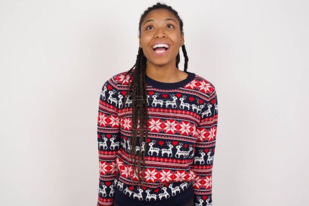 Photo for Surprised African American woman wearing Christmas sweater against white wall shrugs shoulders, looking sideways, being happy and excited. Sudden reactions concept. - Royalty Free Image