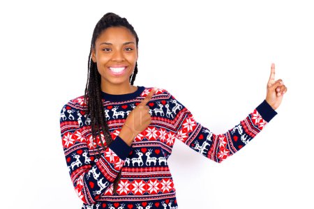 Photo for African American woman wearing Christmas sweater against white wall with positive expression, indicates with fore finger at blank copy space for your promotional text or advertisement. - Royalty Free Image