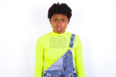 Photo for Portrait of dissatisfied American woman with short hair wearing denim overall against white wall smirks face, purses lips and looks with annoyance at camera, discontent hearing something unpleasant - Royalty Free Image