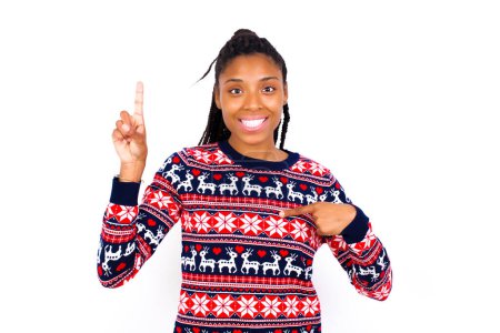 Foto de Mujer afroamericana vistiendo suéter de Navidad contra la pared blanca dice: wow lo emocionante que es, tiene expresión asombrada, indica algo. Una mano en su pecho y señalando con otra mano. - Imagen libre de derechos