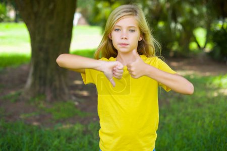 Photo for Caucasian little girl wearing yellow t-shirt standing outdoors showing thumb up down sign - Royalty Free Image