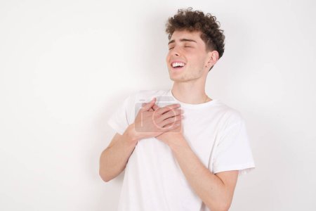 Foto de Joven guapo sobre fondo blanco expresa felicidad, ríe agradablemente, mantiene las manos en el corazón - Imagen libre de derechos