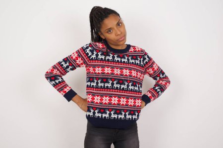 Photo for Funny frustrated African American woman wearing Christmas sweater against white wall holding hands on waist and silly looking at awkward situation. - Royalty Free Image