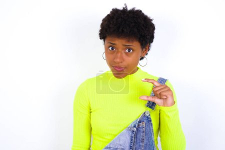Photo for Displeased African American woman with short hair wearing denim overall against white wall shapes little hand sign demonstrates something not very big. Body language concept. - Royalty Free Image