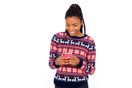 Téléchargez les photos : Femme afro-américaine souriante portant un pull de Noël contre un mur blanc en utilisant un téléphone portable, la messagerie, être heureuse de texter avec des amis, regarder un smartphone. Technologies modernes et communication. - en image libre de droit