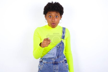 Photo for Shocked American woman with short hair wearing denim overall against white wall points at you with stunned expression - Royalty Free Image