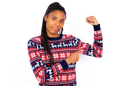 Téléchargez les photos : Femme afro-américaine souriante portant un pull de Noël contre un mur blanc lève la main pour montrer les muscles, se sent confiante dans la victoire, forte et indépendante. - en image libre de droit