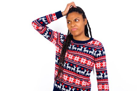 Téléchargez les photos : Femme afro-américaine portant un pull de Noël contre un mur blanc disant : Oups, qu'est-ce que j'ai fait ? Tenant la main sur la tête avec une expression effrayée et de regret. - en image libre de droit