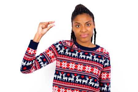 Photo for Upset African American woman wearing Christmas sweater against white wall shapes little gesture with hand demonstrates something very tiny small size. Not very much - Royalty Free Image