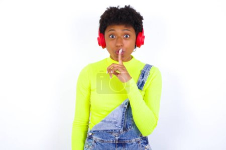 Photo for American woman with short hair wearing denim overall against white wall making hush gesture with finger on her lips wearing  wireless headphones. Be quiet. - Royalty Free Image