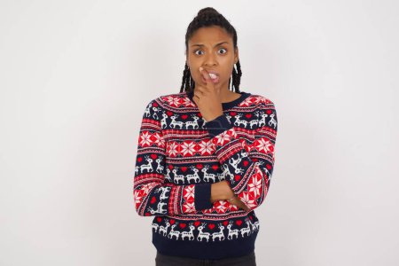 Foto de Mujer afroamericana vistiendo suéter de Navidad contra pared blanca cubriendo la boca con las manos asustadas de algo o alguien mordiendo clavos - Imagen libre de derechos