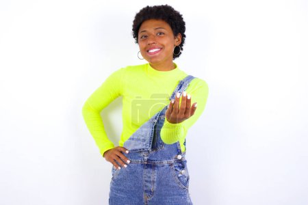 Photo for American woman with short hair wearing denim overall against white wall , inviting you to come, confident and smiling making a gesture with hand, being positive and friendly. - Royalty Free Image