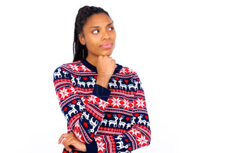 Foto de Mujer afroamericana vistiendo suéter de Navidad contra pared blanca con la mano bajo la barbilla y mirando hacia los lados con expresión dudosa y escéptica, sospechoso y duda. - Imagen libre de derechos