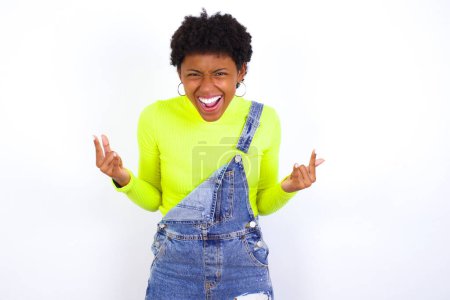 Photo for Born to rock this world. Joyful American woman with short hair wearing denim overall against white wall screaming out loud and showing with raised arms horns or rock gesture. - Royalty Free Image