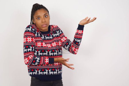 Foto de Mujer afroamericana vistiendo suéter de Navidad contra pared blanca señalando a un lado con ambas manos mostrando algo extraño y diciendo: No sé qué es esto. Concepto de publicidad. - Imagen libre de derechos