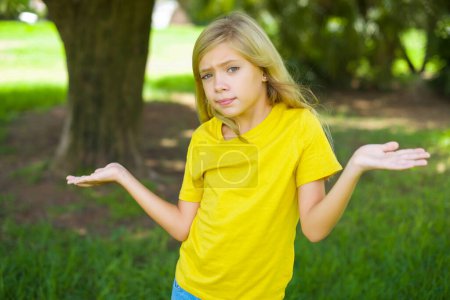 Téléchargez les photos : Insouciante jolie petite fille caucasienne portant t-shirt jaune debout à l'extérieur haussant les épaules, oups. - en image libre de droit