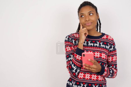 Téléchargez les photos : Pensant femme afro-américaine rêveuse portant un pull de Noël contre un mur blanc à l'aide d'un téléphone portable et tenant la main sur le visage. Prendre des décisions et concept de médias sociaux. - en image libre de droit