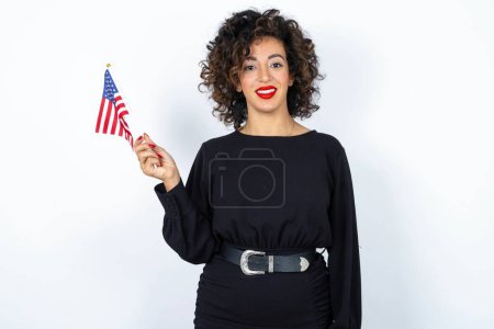 Téléchargez les photos : Jeune belle femme aux cheveux bouclés portant une robe noire et tenant et drapeau américain des États-Unis et souriant sur fond de studio blanc. - en image libre de droit