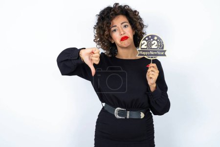 Young beautiful woman with curly hair wearing black dress over white studio background holding a Happy New year 2024 banner and holding thumb down